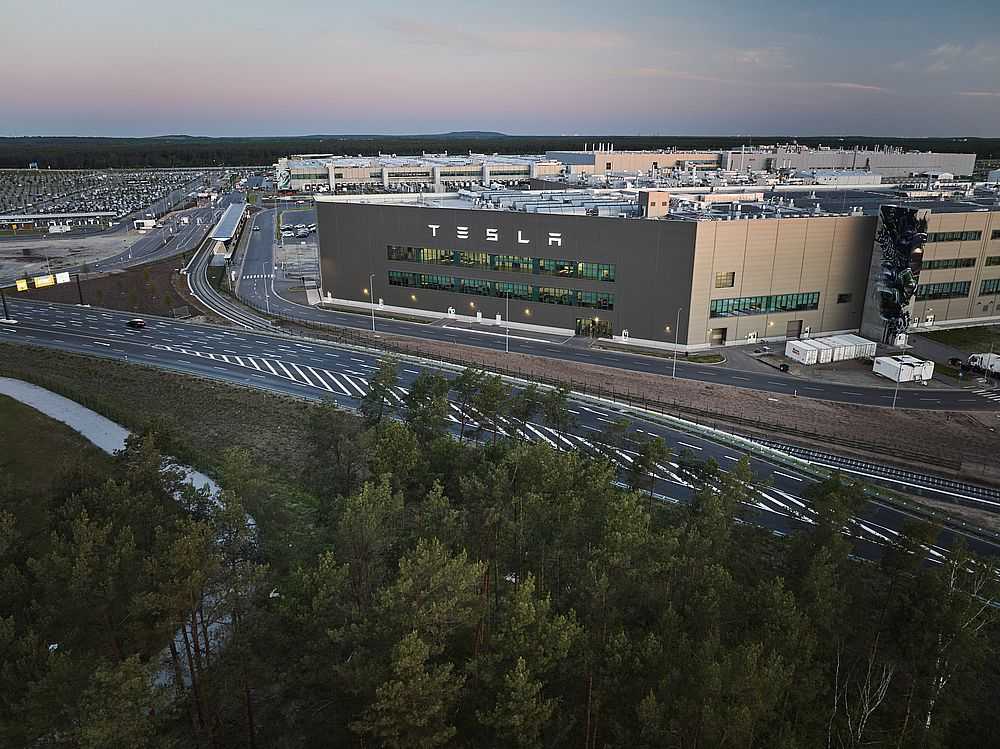 Blick am frühen Morgen auf ein kleines Waldstück im Vordergrund und dem Tesla-Werk im Hintergrund. Tesla-Bahnshuttle zwischen Erkner und Grünheide ist nun in Betrieb - und nicht nur für Mitarbeiter praktisch. (Foto: Jörg Carstensen/dpa)